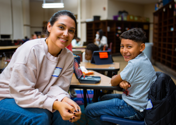 A tutor (woman) student (young boy) pair sit at a table facing the camera smiling while taking a break from their tutoring session. They are seated at a desk in a classroom. Other tutoring chicago students and tutors are present in the background of the image.