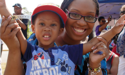 A young boy wearing a PAW patrol shirt and a baseball cap stands smiling with his mom behind him. 